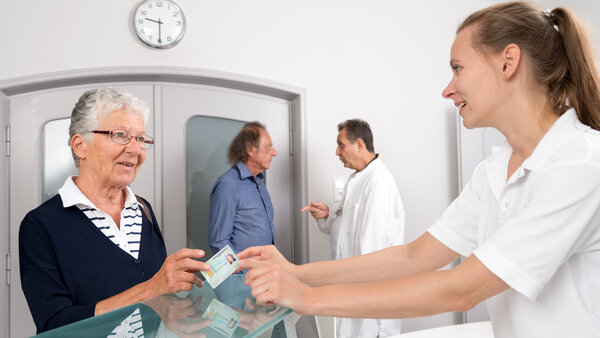Arztpraxis: Empfang, © Krebsinformationsdienst, DKFZ; Foto: Tobias Schwerdt Patientin reicht einer Arzthelferin ihre Versichertenkarte am Empfang einer Arztpraxis. Im Hintergrund dpricht ein Arzt mit einem Patienten.