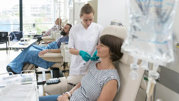 Chemoinfusin vorbereiten, © Krebsinformationsdienst, DKFZ; Foto: Tobias Schwerdt Eine Patientin sitzt in einer Chemo-Ambulanz mit anderen Patienten. Eine Krankenschwester bereitet ihre Infusion vor.