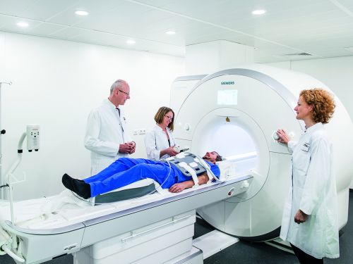 Ein Patient liegt auf der Liege eines Kernspinntomographen, während Ärzte neben ihm stehen und den Vorgang erklären.