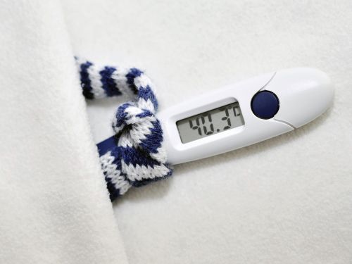 Ein Fieberthermometer, das 40,3 Grad anzeigt, mit einem winzigen blau-weiß gestreiften Schal liegt in einem winzigen weißen Bett.