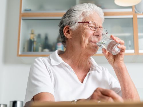 Ältere Frau sitzt in der Küche und trinkt Wasser aus einem Glas.