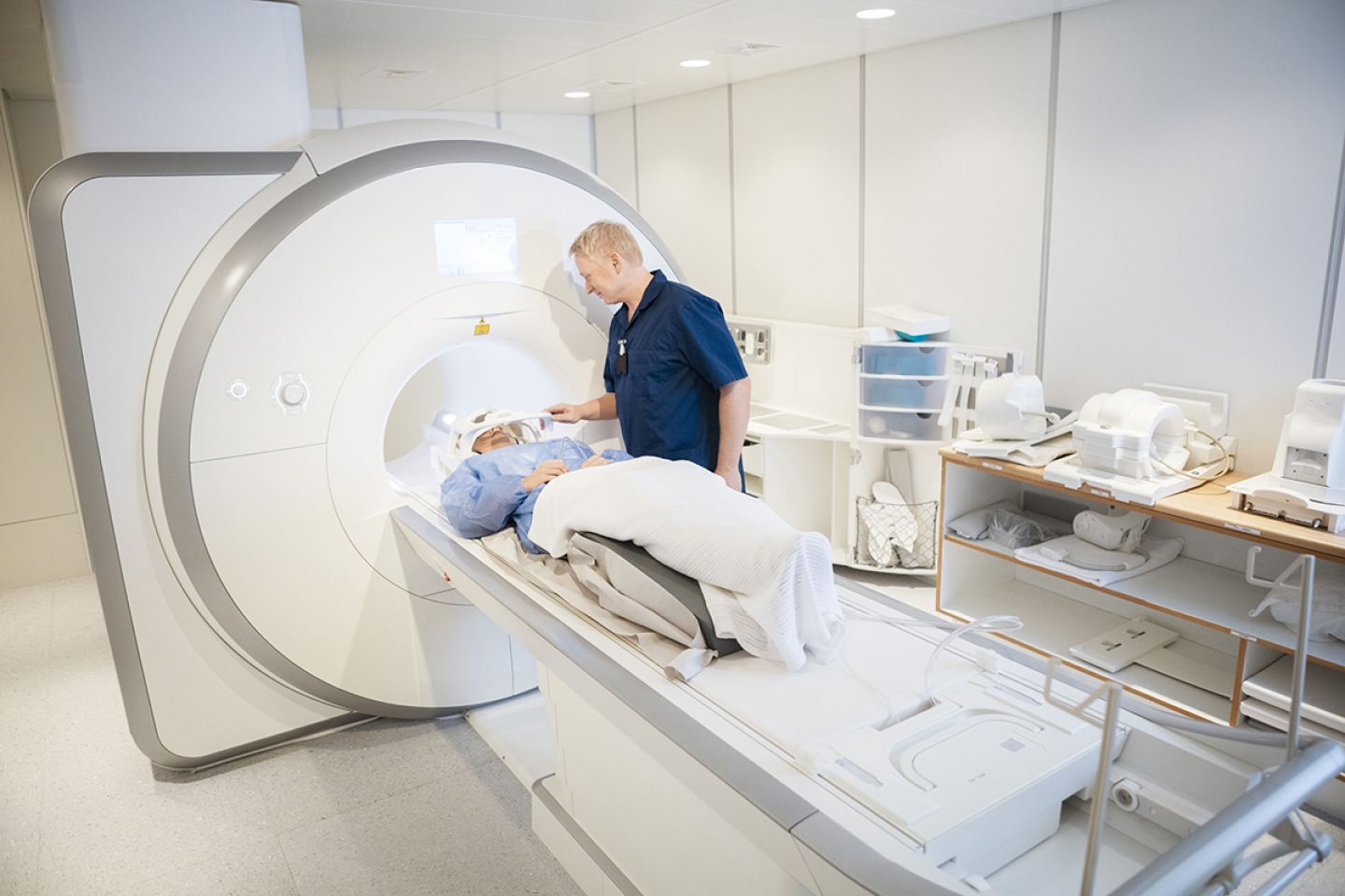 Eine Frau liegt auf dem Untersuchungstisch eines Magnetresonanztomographen, neben ihr steht ein Mann in einem blauen Kittel.