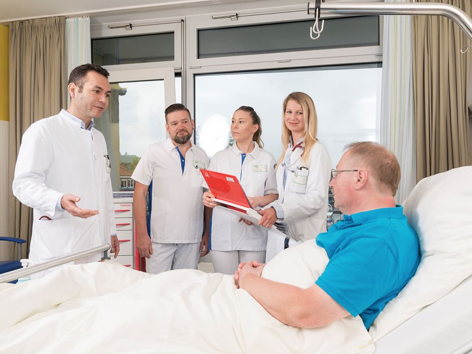 Ärzte und Pflegende stehen bei der Visite um einen im Bett liegenden Patienten und sprechen mit ihm.
