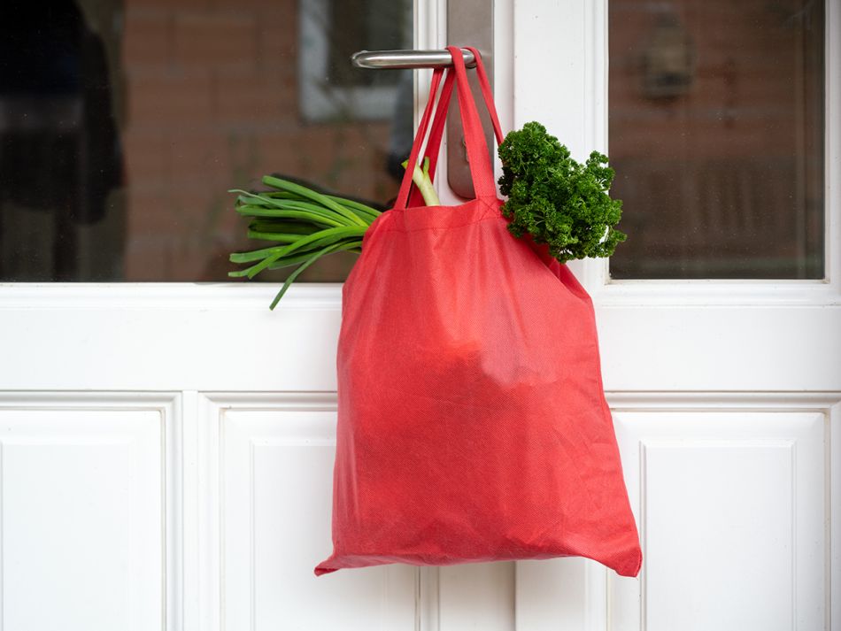 Rote Einkaufstasche mit Gemüse hängt außen an einer weißen Haustüre.