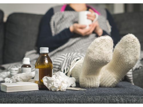 Frau mit Erkältung sitzt auf einem Sofa umgeben von Taschentüchern und Medikamenten..