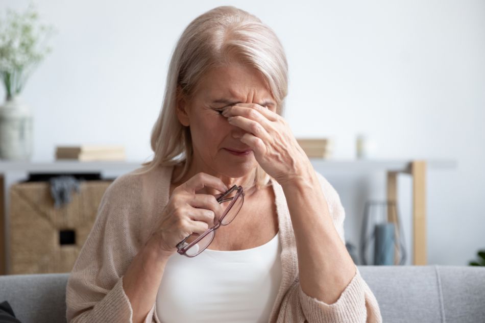 Ältere Frau mit schulterlangen grauen Haaren fasst sich mit schmerzverzerrtem Gesicht mit der linken Hand an die Stirn, während sie eine rote Brille in der rechten Hand hält.