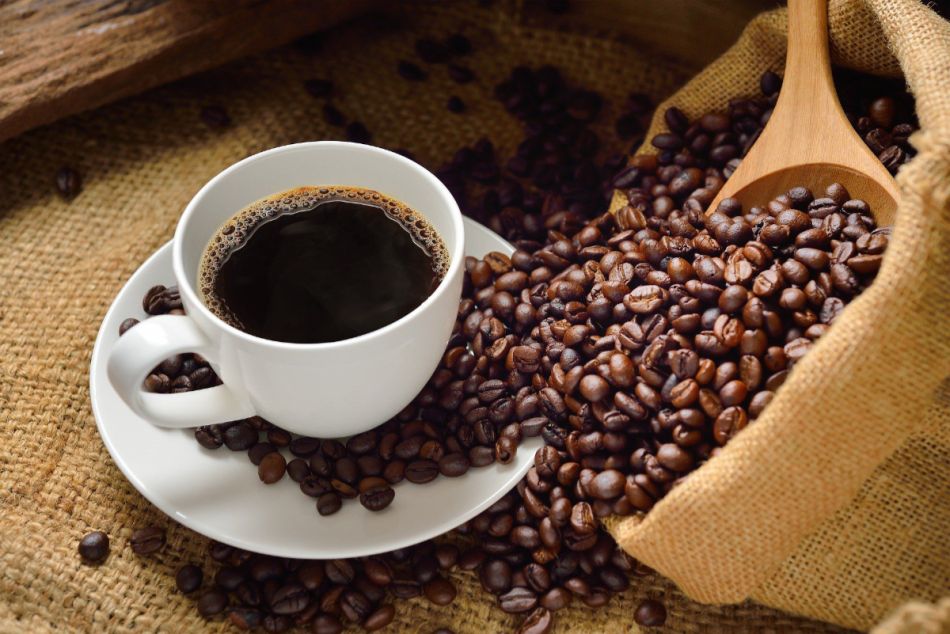 Eine Tasse schwarzer Kaffee steht auf einem Tisch. Rechts daneben ist ein Jutebeutel mit Kaffeebohnen und einem Holzlöffel.