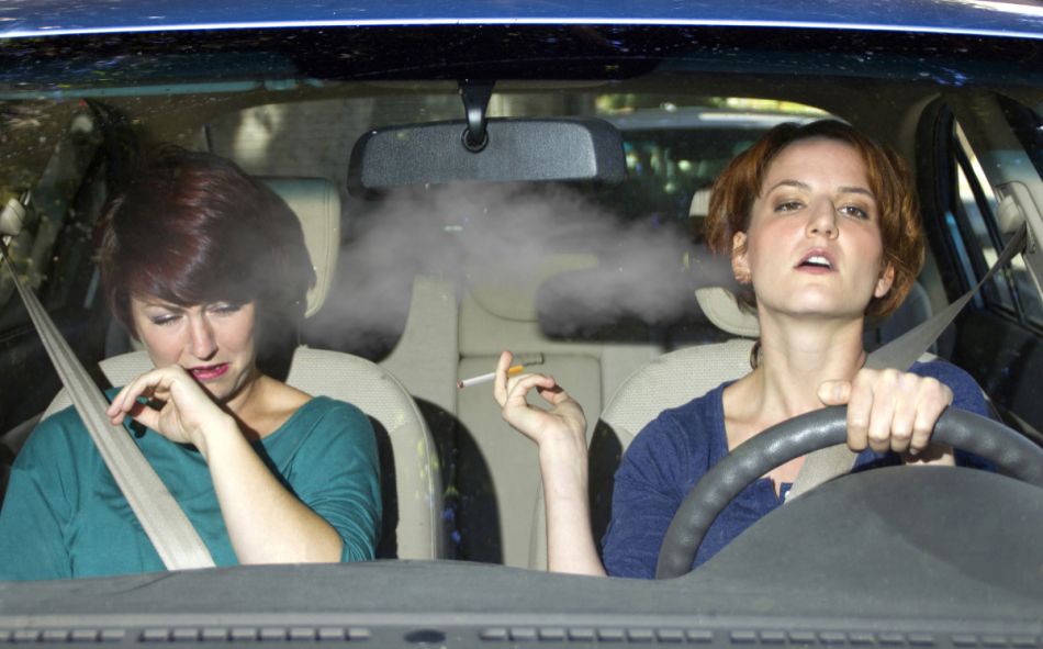 Eine junge Raucherin sitz hinter dem Steuer eines Autos - die Beifahrerin wendet sich hustend ab