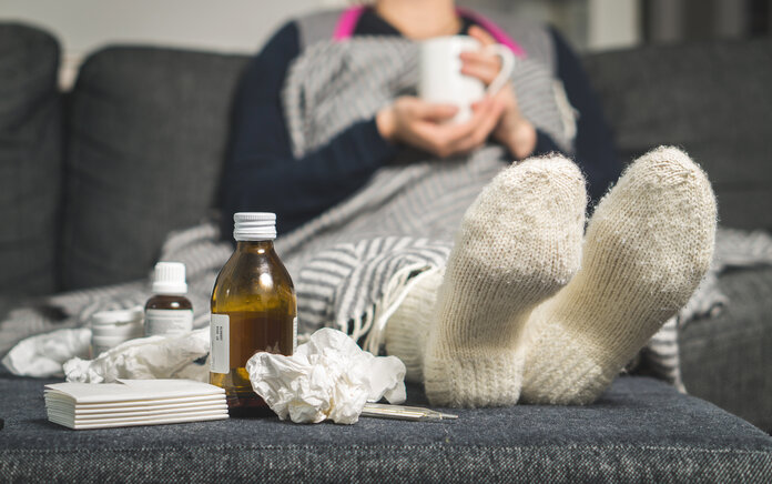 Im Bildvordergrund sind Medikamente und Taschentücher zu sehen; im Bildhintergrund liegt eine zugedeckte Frau auf einem Sofa, die eine Tasse mit beiden Händen hält.