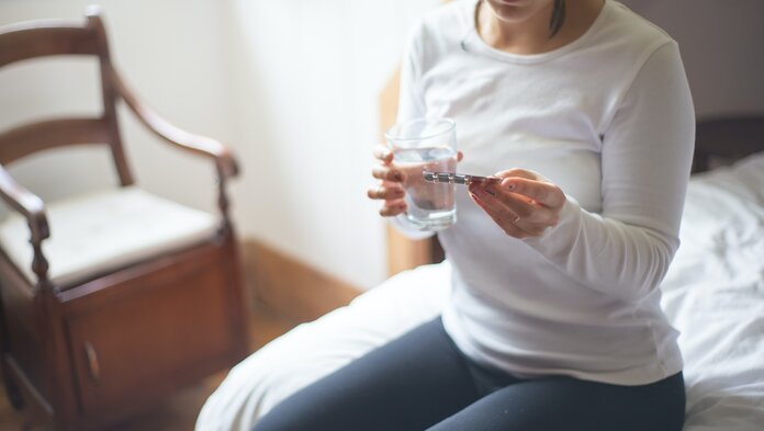 Junge Frau sitzt auf dem Bett mit einem Tablettenblister in der einen und einem Glas Wasser in der anderen Hand.