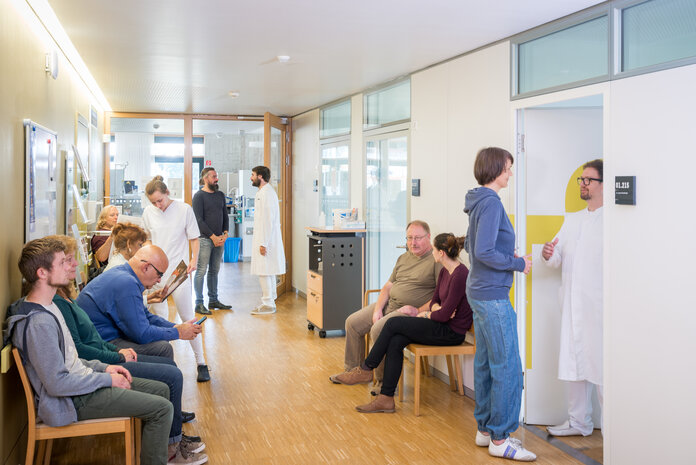 Mehrere Patienten und Patientinnen sitzen im Wartezimmer einer Tagesklinik oder Chemo-Ambulanz. Ärzte stehen im Flur und in der Tür zum Behandlungszimmer.