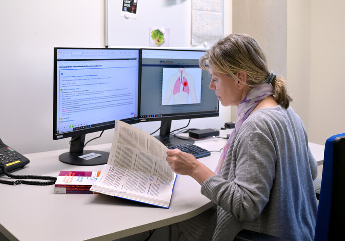 Eine Mitarbeiterin vom Wissensmanagement des Krebsinformationsdienstes sitzt am PC, blättert in einem lehrbuch und arbeitet.