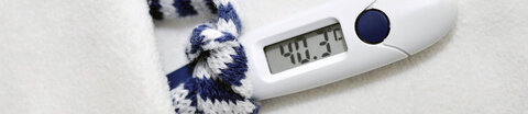 Fieberthermometer, das 40,3 Grad anzeigt, einen winzigen blau-weiß gestreiften Schal trägt und in einem winzigen weißen Bett liegt