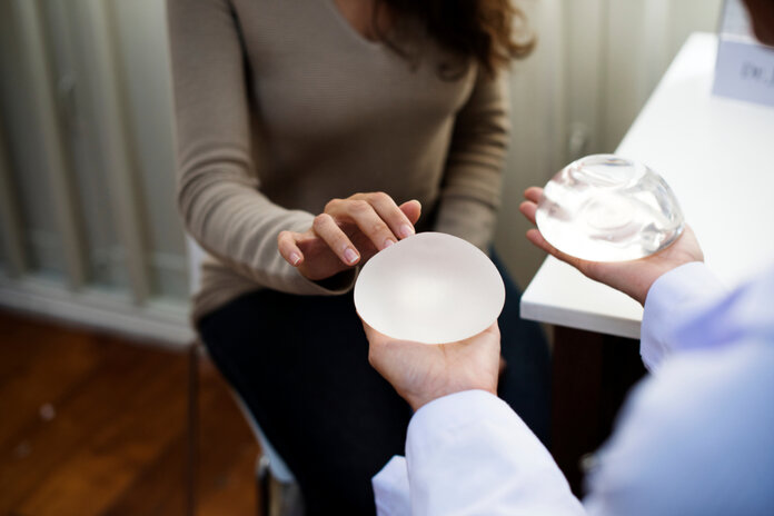 Arzt hält ein Brustimplantat mit glatter und ein Implantat mit aufgerauter Oberfläche in der Hand, während eine Frau das linke Implantat berührt. © Rawpixel.com, Shutterstock