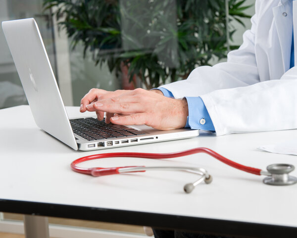 Die Hände eines Arztes tippen auf der Tastatur eines Laptops, daneben liegt ein Stethoskop.