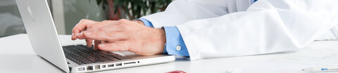 Die Hände eines Arztes tippen auf der Tastatur eines Laptops, daneben liegt ein Stethoskop.