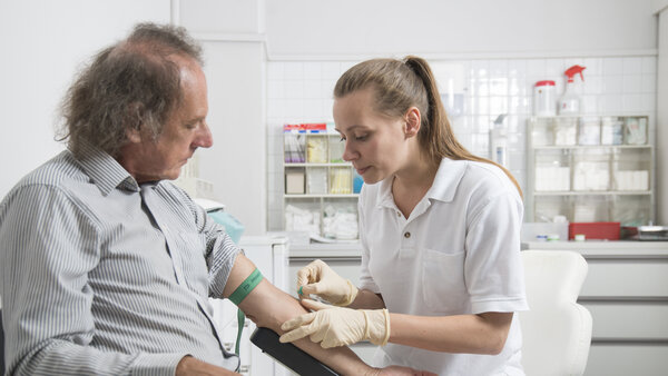 Ein Mann sitzt auf einem Stuhl in der Arztpraxis, während eine Arzthelferin Blut abnimmt.