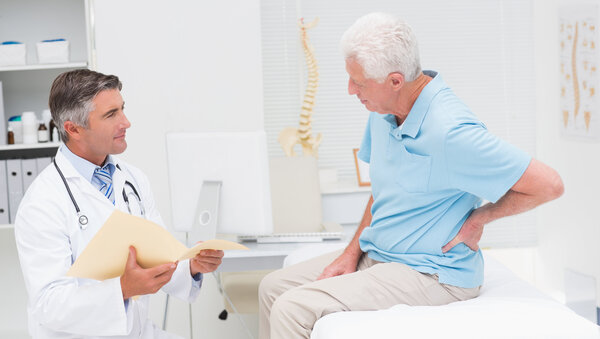 Arztgespräch zu Rückenschmerzen, © ESB Professional, Shutterstock Ein Arzt sitzt mit einer aufgeklappten Patientenakte vor einem älteren Patienten, der auf einer Patientenliege sitzt und sich den Rücken hält.