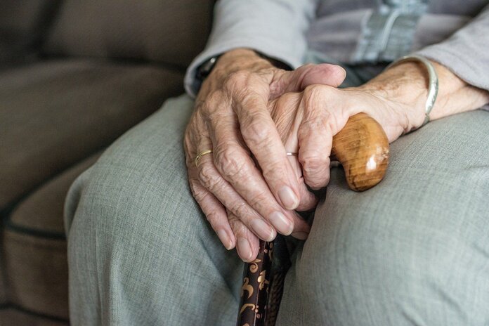 Nahaufnahme einer Seniorin, die mit übereinander gelegten Händen auf dem Sofa sitzt.