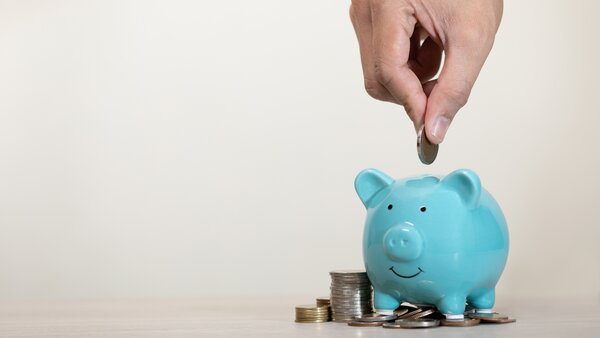 Spendenschwein, © Natthapol Siridech, Shutterstock Ein Sparschwein steht vor einem Stapel Münzen; von oben steckt eine Hand ein Geldstück in den Schlitz