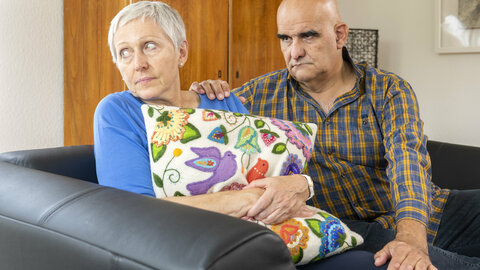 Ein älteres Paar sitzt auf dem Sofa. Die Frau hält ein Kissen an sich gedrückt und schaut traurig weg. Der Mann hat tröstend die Hand auf die Schulter der Frau gelegt und schaut betreten.