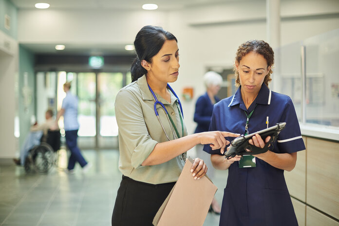 Eine Ärztin und eine Pflegerin stehen im Klinikflur und schauen gemeinsam auf ein Tablet. Die Ärztin hält ausgedruckte Dokumente in einer Hand.