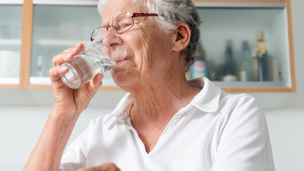 Ältere Frau sitzt in der Küche und trinkt Wasser aus einem Glas.