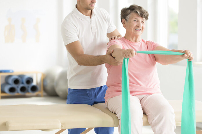 Ein Physiotherapeut unterstützt eine Seniorin bei Übungen mit dem Terraband.