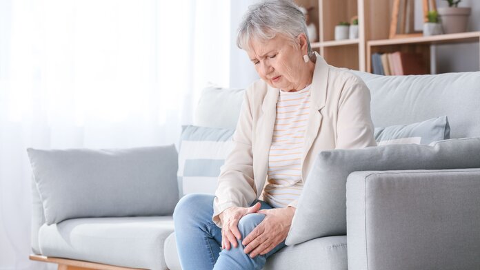 Ältere Frau sitzt auf dem Sofa und umfasst schmerzerfüllt ihr Knie.