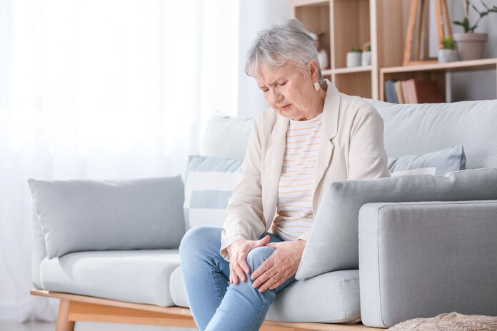 Ältere Frau sitzt auf dem Sofa und umfasst schmerzerfüllt ihr Knie.