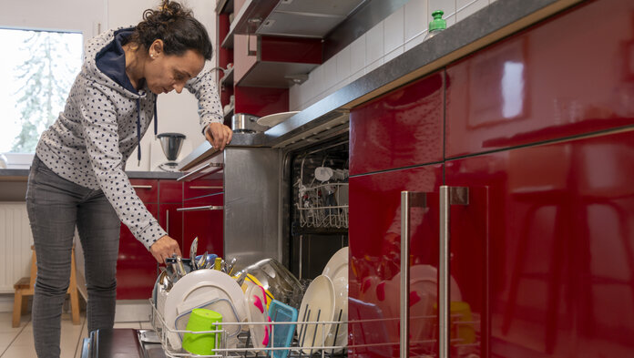Eine Frau mittleren Alters räumt die Spülmaschine aus und stützt sich dabei auf der Arbeitsplatte ab.