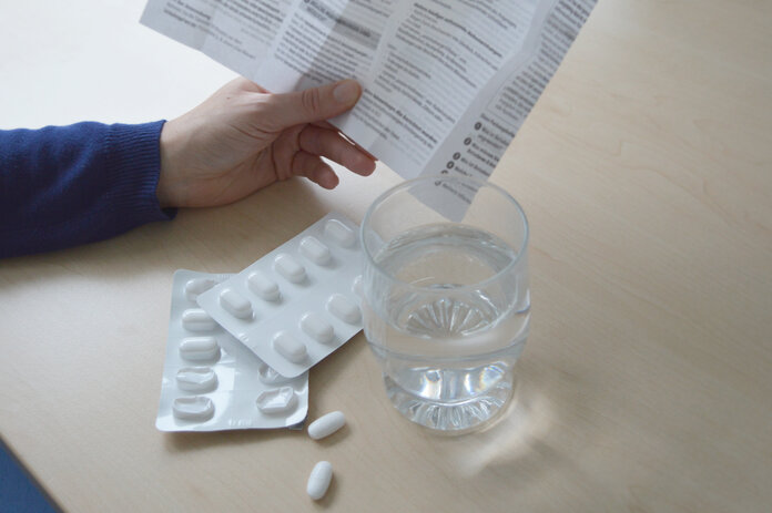 Eine Hand hält eine Packungsbeilage, davor liegen Tabletten auf den Tisch sowie ein Glas Wasser.