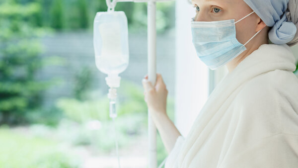 Patientin mit Kopftuch und Mundschutz hält einen Infusionsständer und schaut aus dem Fenster.