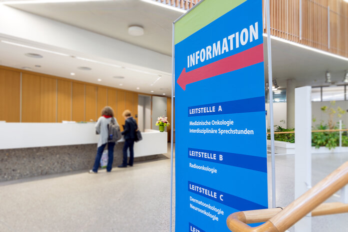 Eingangsbereich Krankenhaus mit Informationstafel und Personen.