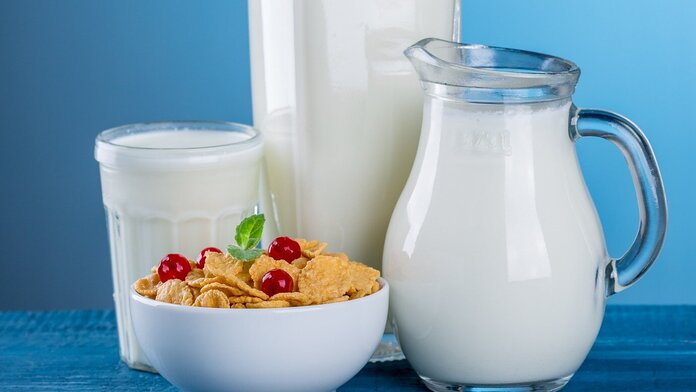 Ein Glas, eine Flasche und eine Karaffe mit Milch stehen zusammen mit einer Schale Cerealien auf einem Tisch.