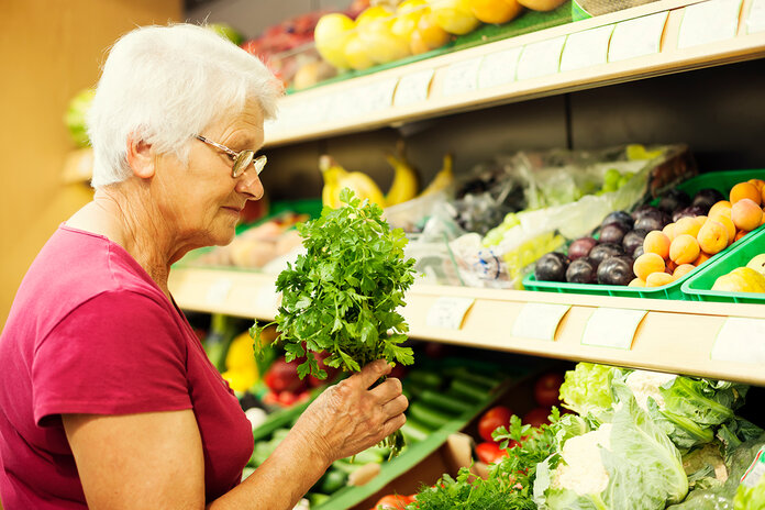Seniorin ist im Supermarkt in der Obst- und Gemüseabteilung und hält Petersilie in der Hand.