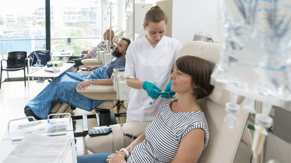 Eine Patientin sitzt in einer Chemo-Ambulanz mit anderen Patienten. Eine Krankenschwester bereitet ihre Infusion vor.