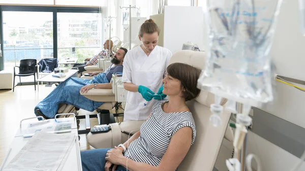 Chemoinfusin vorbereiten, © Krebsinformationsdienst, DKFZ; Foto: Tobias Schwerdt Eine Patientin sitzt in einer Chemo-Ambulanz mit anderen Patienten. Eine Krankenschwester bereitet ihre Infusion vor.