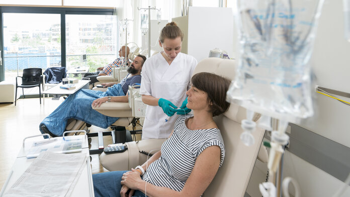 Eine Patientin sitzt in einer Chemo-Ambulanz mit anderen Patienten. Eine Krankenschwester bereitet ihre Infusion vor.