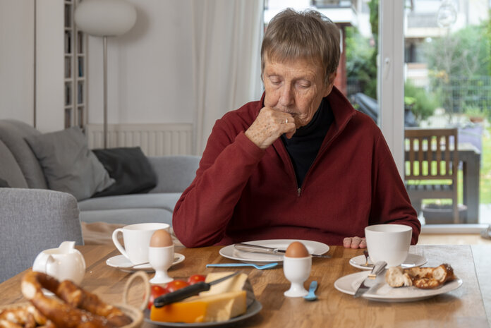 Eine ältere Frau sitzt ohne Appetit an einem Esstisch mit Frühstück.