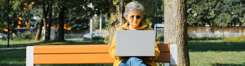 Eine ältere Frau sitzt mit ihrem Laptop auf einer Parkbank.