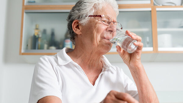 Ältere Frau sitzt in der Küche und trinkt Wasser aus einem Glas.