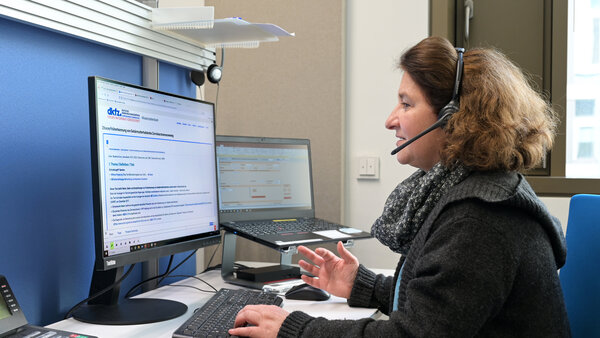 Eine Ärztin vom Telefondienst des Krebsinformationsdienstes sitzt an einem Arbeitsplatz mit 2 Computer-Bildschirmen, auf die sie schaut, während sie über ein Headset telefoniert.