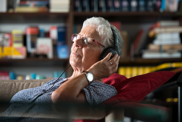 Ältere Frau beim Musikhören mit Kopfhörer auf einem Sofa.