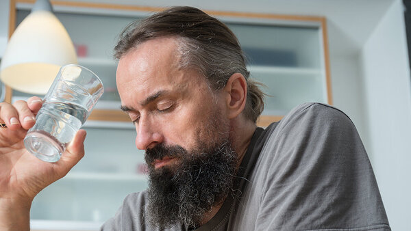 Ein Mann sitzt mit geschlossenen Augen und einem Wasserglas am Tisch.