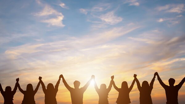 Gemeinschaft, © Guitarfoto studio, Shutterstock Man sieht die Silhouette einer Menschengruppe vor einem Sonnenuntergang die ihre Arme hoch halten und sich an den Händenfassen.