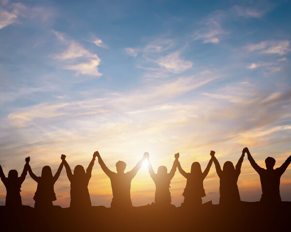 Man sieht die Silhouette einer Menschengruppe vor einem Sonnenuntergang die ihre Arme hoch halten und sich an den Händenfassen.