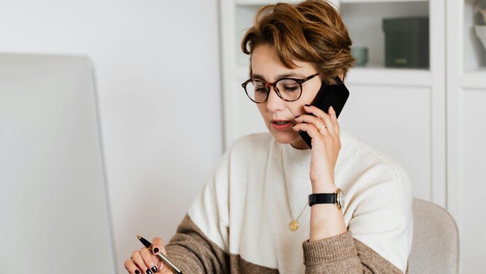 Eine Frau sitzt am Computer und telefoniert mit ihrem Handy am Ohr.