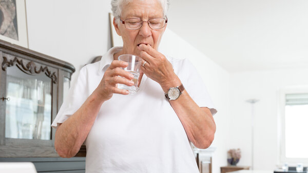 Ältere Frau nimmt Medikamente mit einem Glas Wasser ein. Vor ihr auf dem Tisch liegt eine Dosierhilfe.