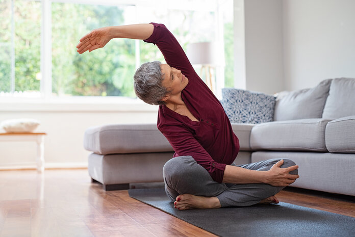 Ältere Frau beim Yoga im Wohnzimmer., © Rido, Shutterstock Ältere Frau macht eine Dehnübung auf einer Yogamatte neben einem Sofa.